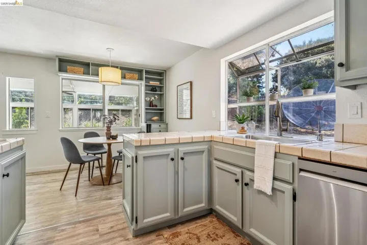 Kitchen featuring light gray cabinetry with black hardware, tile countertops, stainless steel dishwasher, and a garden window above the sink
