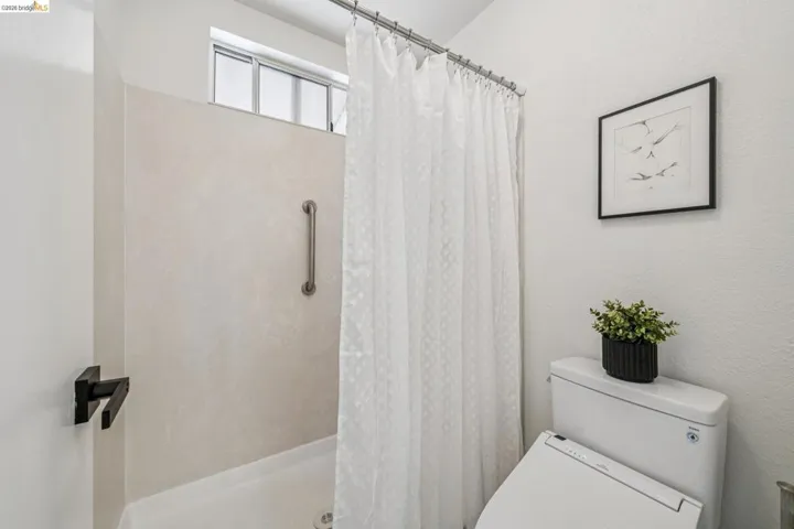 Shower stall with a grab bar, featuring a transom window, white shower curtain, and a white toilet