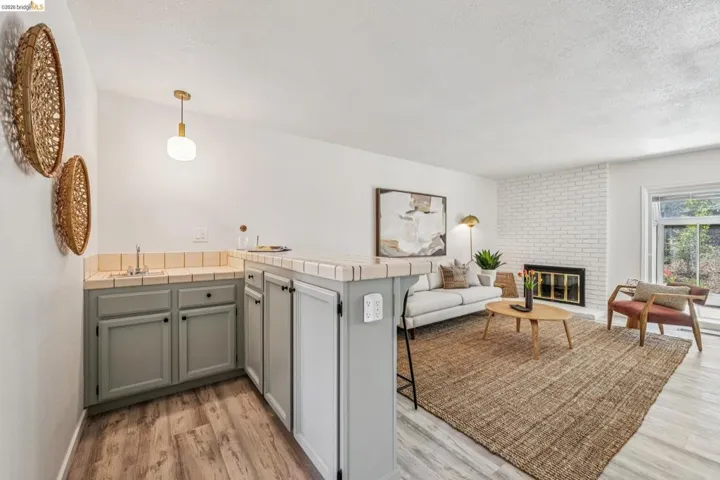 Living area featuring a white brick fireplace, wood-finish flooring, and a wet bar with light gray cabinetry and a tile countertop