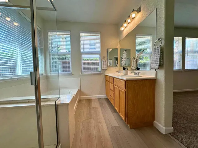 Bathroom featuring a double vanity with wood cabinetry, a built-in tub, a separate shower with a glass enclosure, wood-finish flooring, and multiple windows with blinds