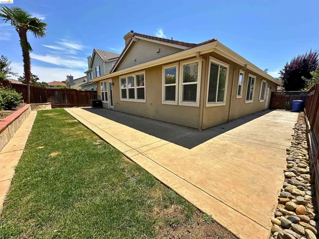 Exterior stucco finish featuring numerous windows, a concrete patio, a grass lawn, and a retaining wall with stone cap
