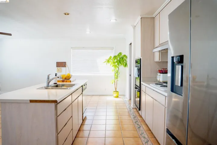Kitchen featuring a central island with an integrated sink, light-toned cabinetry, stainless steel appliances including a refrigerator and double wall ovens, and terracotta-style floor tiling