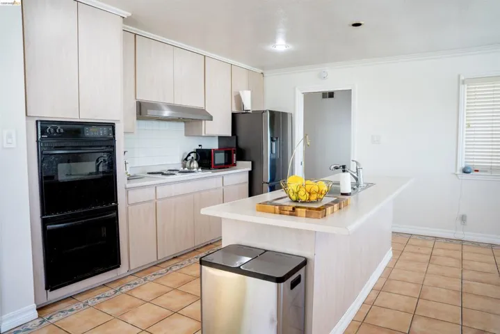 Kitchen featuring an integrated double oven, light wood-finish cabinetry, a white subway tile backsplash, a center island with sink, and terracotta-style floor tiles