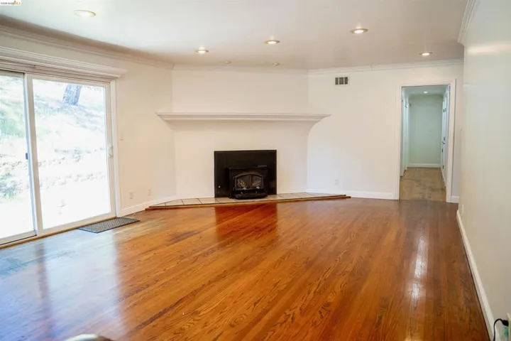 Spacious living area featuring rich wood-finish flooring, a prominent fireplace with a white mantel, recessed lighting, white baseboards, and a large glass sliding door opening to exterior surroundings