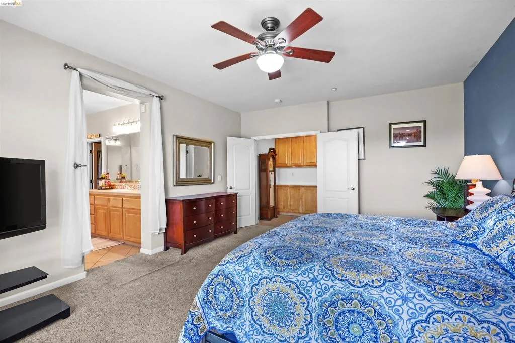 Carpeted bedroom featuring a ceiling fan with integrated lighting, a blue accent wall, and a doorway leading to a bathroom with dual vanities and a tiled floor