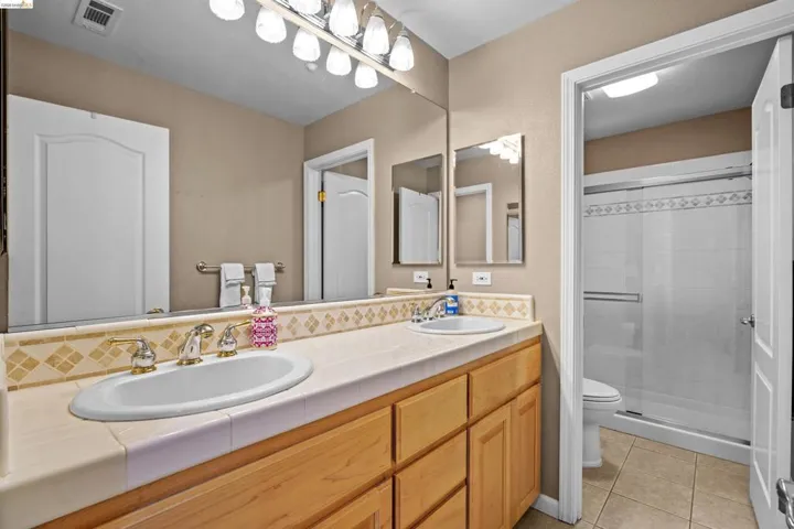 Double vanity with light wood cabinetry, tiled countertop, and a full-width mirror