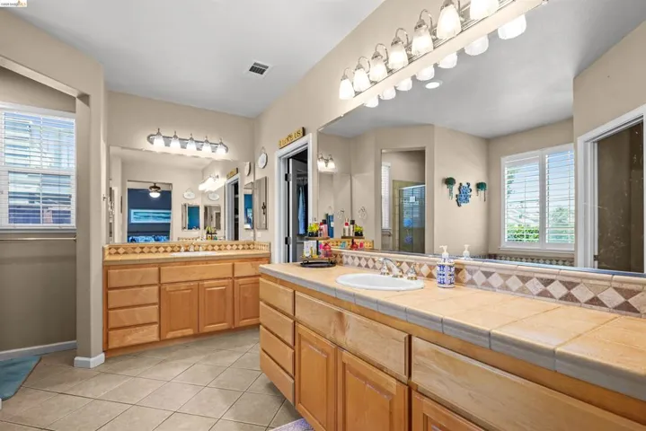 Bathroom vanity with dual sinks, ceramic tile countertops, light wood-finish cabinetry, and tile flooring