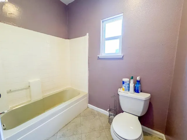 Bathroom featuring a white bathtub with subway tile surround, neutral-toned floor tiling, a white toilet, and a window