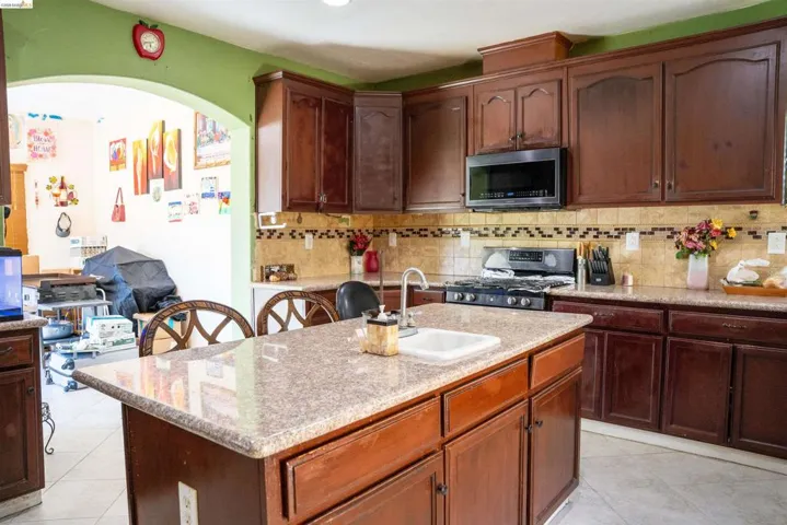Kitchen featuring dark wood cabinetry, a central island with an integrated sink, a gas range with an overhead microwave, tile countertops, and a decorative tile backsplash