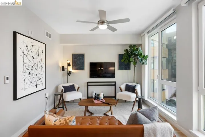 Spacious living area featuring light-toned walls, wood-finish flooring, a contemporary ceiling fan, and large windows providing abundant natural light