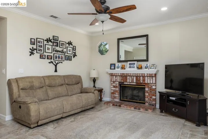 Living space featuring a brick fireplace with a mantel, recessed lighting, crown molding, a ceiling fan with wood-finish blades, and a hexagonal window