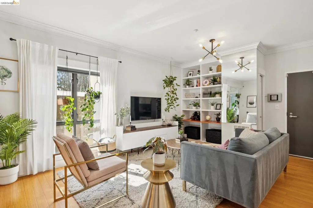 Living space featuring wood-finish flooring, a full-wall built-in shelving unit, a large window with white curtains, a flush-mount light fixture, and crown molding