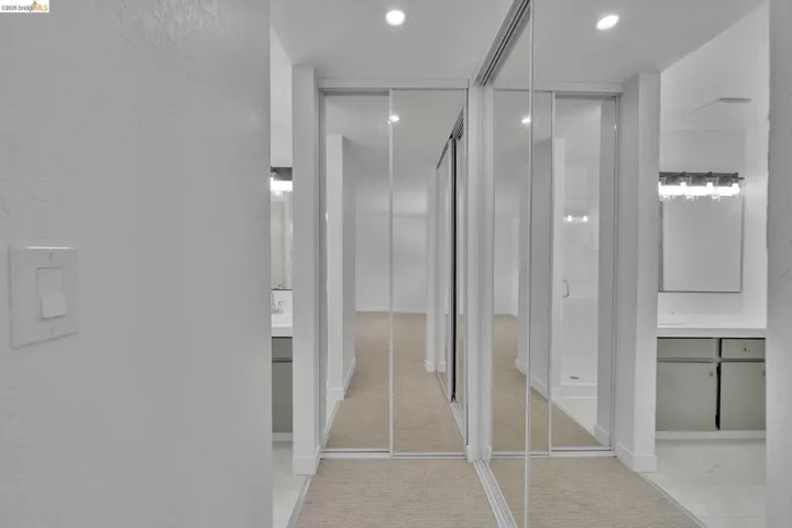 Mirrored sliding closet doors and recessed lighting illuminate a carpeted hallway, flanked by two vanity areas with integrated sinks and sconce lighting