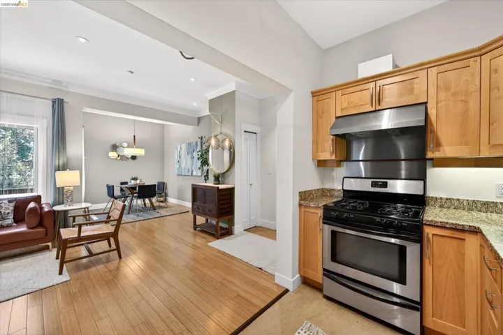 Open-concept kitchen and living area featuring wood-finish flooring, stainless steel range with overhead hood, light wood cabinetry, and granite countertops