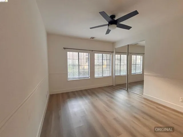 Spacious room featuring wood-finish flooring, two windows with blinds, a ceiling fan with integrated lighting, and mirrored closet doors