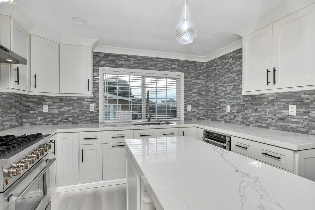 Kitchen featuring stainless steel appliances, white cabinets, light stone counters, decorative light fixtures, and ornamental molding