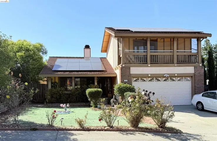 Two-story residence featuring a brick facade, integrated solar panels, an upper-level balcony with a wood railing, an attached two-car garage, and a manicured front lawn