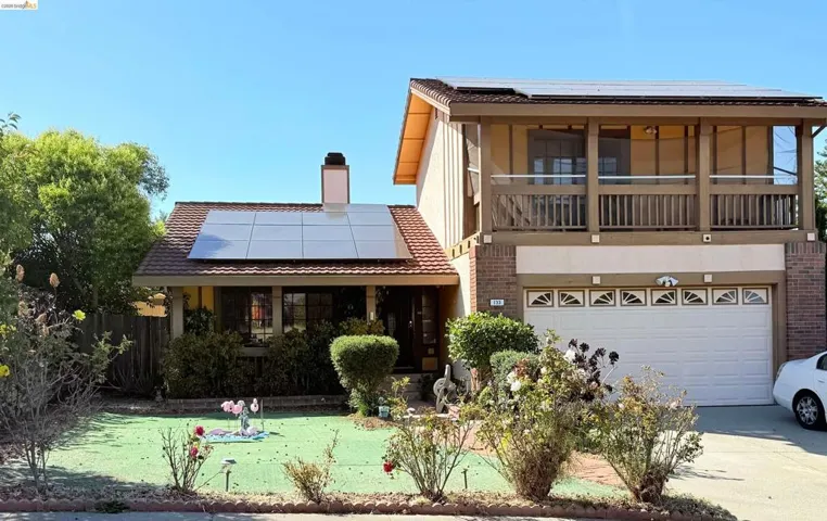 Two-story residence featuring a brick facade, integrated solar panels, an upper-level balcony with a wood railing, an attached two-car garage, and a manicured front lawn