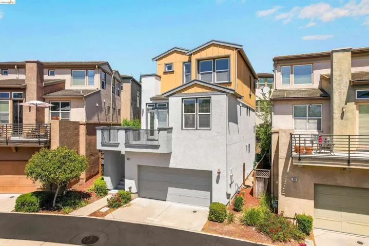 Modern townhouse featuring a light gray stucco exterior with wood-tone accents, an attached garage, and a private balcony with dark gray railings
