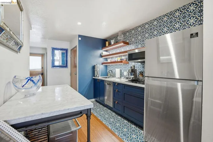 Kitchenette featuring a stainless steel refrigerator, blue patterned floor tile, blue base cabinetry with brass pulls, marble-look countertops, and open wood shelving with under-shelf lighting