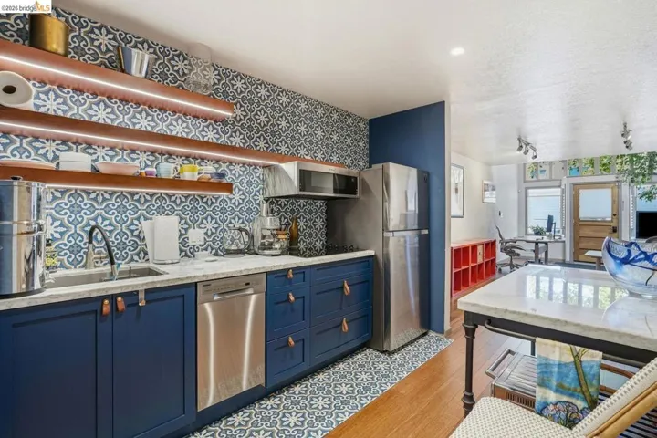Kitchenette featuring blue cabinetry with leather pulls, stainless steel appliances, open wood-finish shelving with integrated lighting, a patterned tile backsplash, and a coordinating patterned floor tile