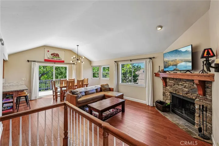 Open-concept Lake Arrowhead living area with vaulted ceilings, warm wood-style flooring, a dramatic stone fireplace, abundant natural light, and easy flow to the dining area and outdoor deck.