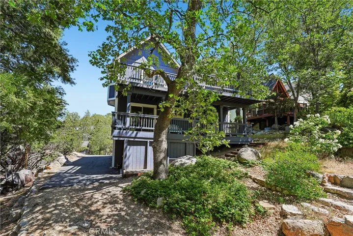 Exterior view from the lower yard showing the home’s elevated deck, natural boulder landscaping, mature trees, and mountain neighborhood setting.