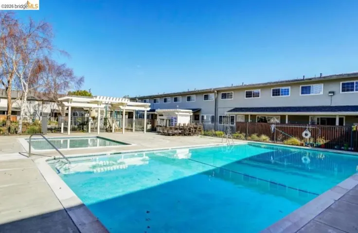 Outdoor swimming pool with concrete surround, featuring a smaller adjacent pool, pergola-covered seating areas, and perimeter fencing