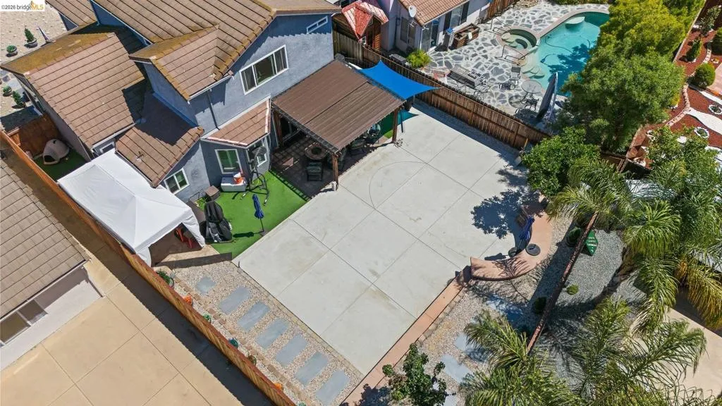 Expansive concrete patio area featuring a metal pergola, artificial turf, perimeter fencing, and mature landscaping