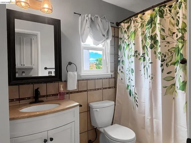 Bathroom featuring a white vanity with a tan countertop, a rectangular mirror with a dark frame, a toilet, and light-toned wall tiling