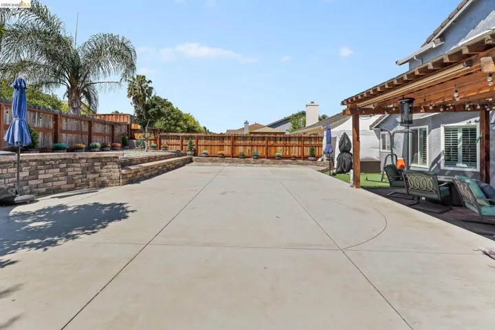 Expansive concrete patio area featuring a wooden pergola with string lighting, a stone-faced retaining wall, and wood privacy fencing