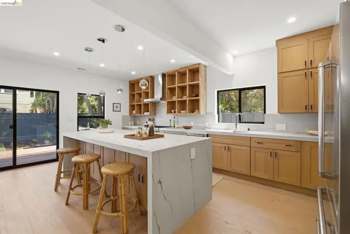 Modern kitchen featuring a waterfall island with stone-finish countertop, light wood-finish cabinetry, built-in shelving, stainless steel appliances, and a sliding glass door leading to a deck