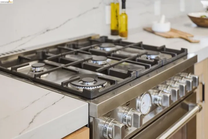 Stainless steel gas range with six burners and heavy-duty grates, integrated into a white countertop with subtle veining, complemented by light wood-tone cabinetry