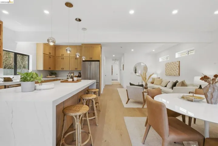 Open-concept living area with light wood-finish flooring, a contemporary kitchen featuring light wood cabinetry and a large island with a waterfall countertop, recessed lighting, and a stainless steel refrigerator