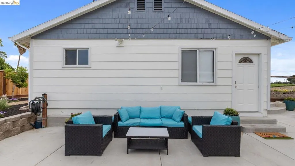 Spacious concrete patio featuring a white siding exterior with a grey shingle gable, a built-in garden bed with stone retaining wall, and an arched panel door