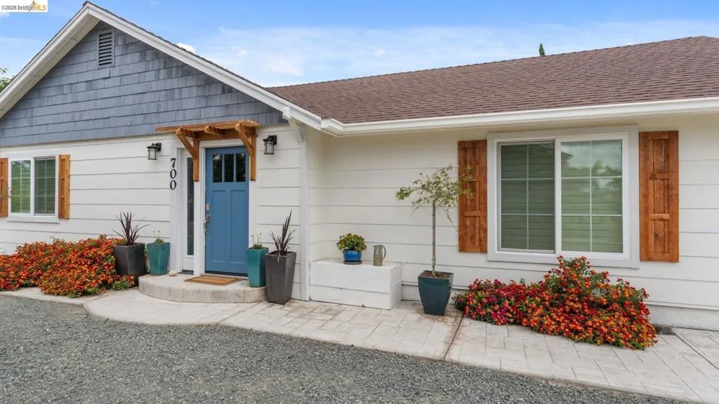 Exterior facade featuring white siding with contrasting blue shingle accents, a blue paneled entry door with an overhead wooden trellis, and white-framed windows with decorative wooden shutters