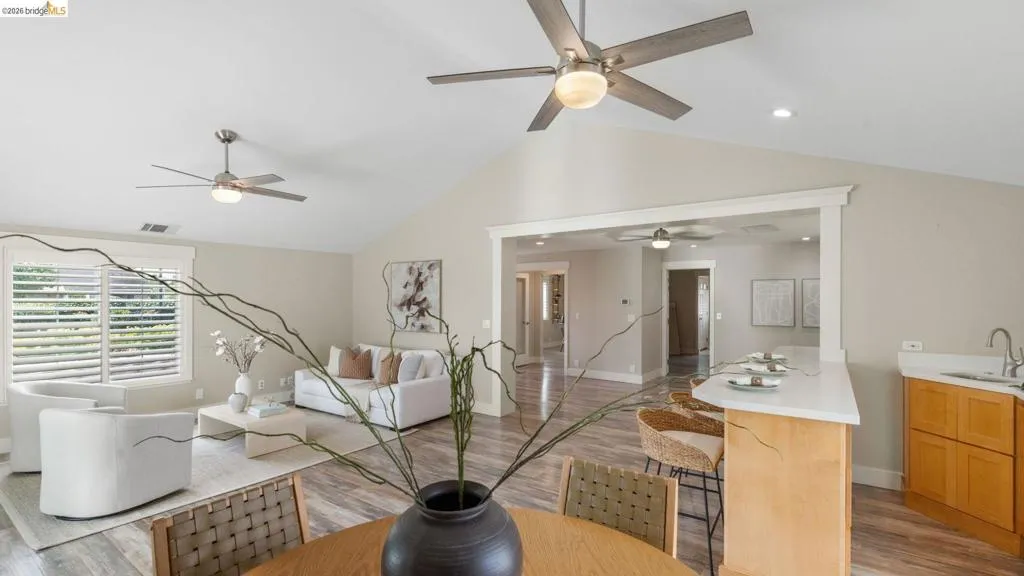 Open-concept living space featuring wood-finish flooring, vaulted ceilings, plantation shutters, a kitchen island with a white countertop, and light wood cabinetry