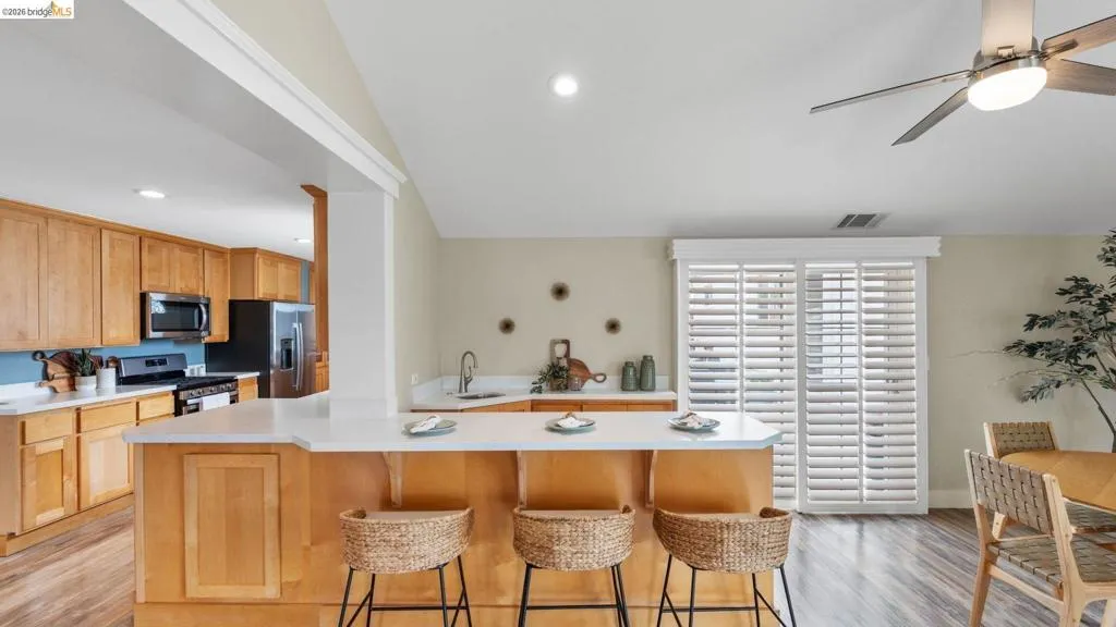 Open-concept kitchen and dining area featuring light wood-finish cabinetry, stainless steel appliances, a breakfast bar with white countertops, and sliding glass doors with plantation shutters