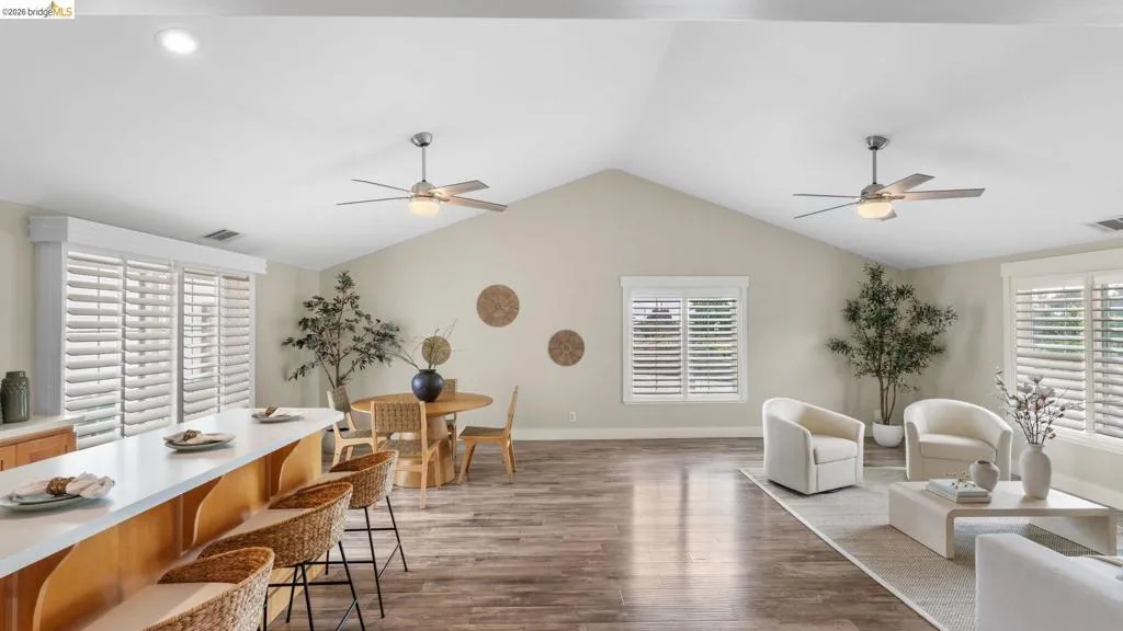 Vaulted ceilings enhance this open-concept living space, featuring wood-finish flooring, two ceiling fans, and multiple windows with plantation shutters