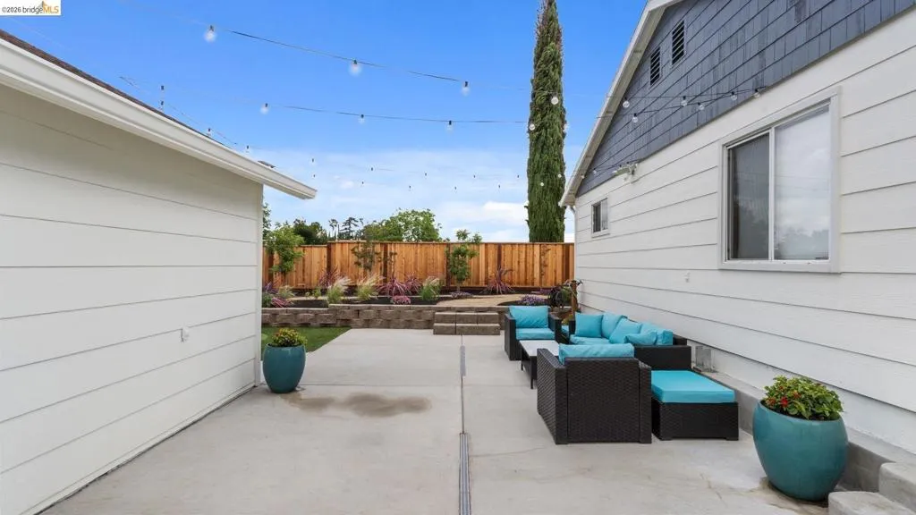 Hardscaped patio with string lighting, multiple tiered garden beds, wooden privacy fencing, and exterior house siding