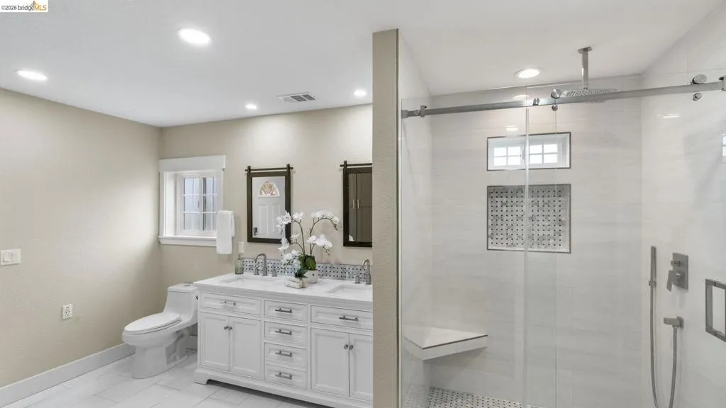Spacious bathroom featuring a dual vanity with white cabinetry and undermount sinks, a window with white trim, a toilet, and a glass-enclosed shower with a built-in bench and recessed niche