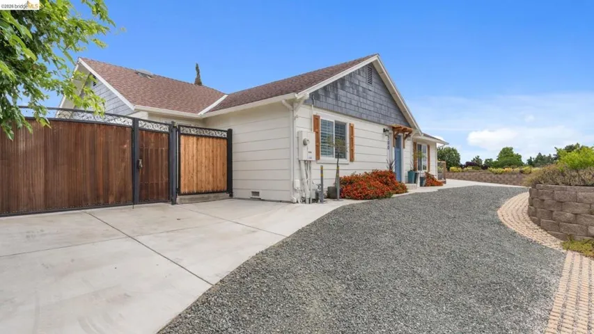 Wood panel and wrought iron gates, gravel driveway, concrete pad, horizontal siding exterior, and a stone retaining wall