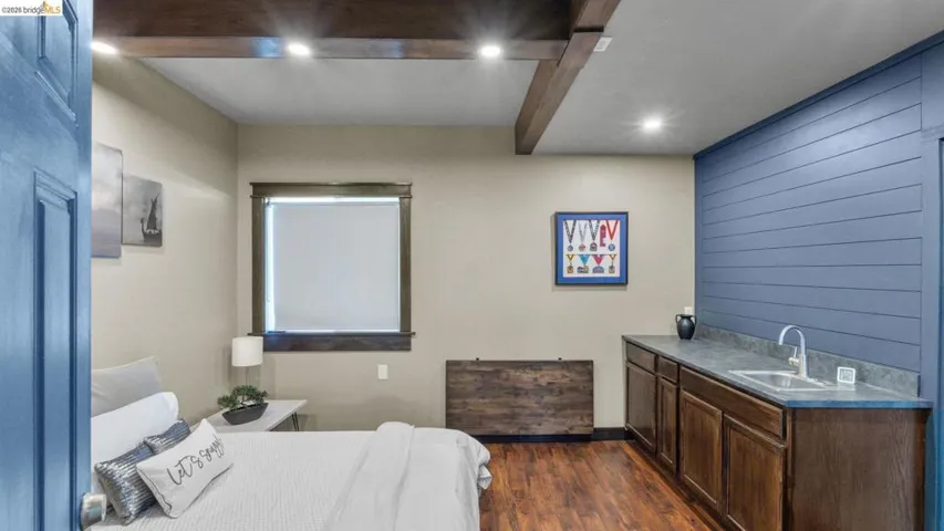 Interior room featuring wood-finish flooring, exposed ceiling beams with recessed lighting, and a window with a roller shade