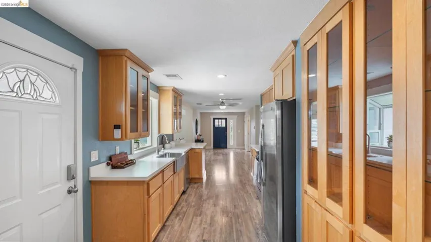 Open-concept kitchen featuring wood-finish flooring, light wood cabinetry, white countertops, stainless steel appliances, and a farmhouse sink