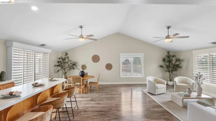 Vaulted ceilings enhance this open-concept living space, featuring wood-finish flooring, two ceiling fans, and multiple windows with plantation shutters