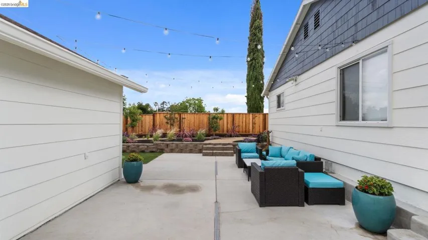 Hardscaped patio with string lighting, multiple tiered garden beds, wooden privacy fencing, and exterior house siding
