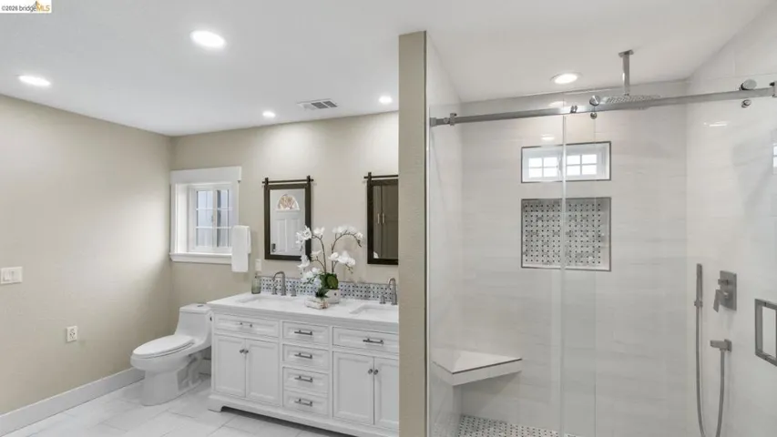 Spacious bathroom featuring a dual vanity with white cabinetry and undermount sinks, a window with white trim, a toilet, and a glass-enclosed shower with a built-in bench and recessed niche
