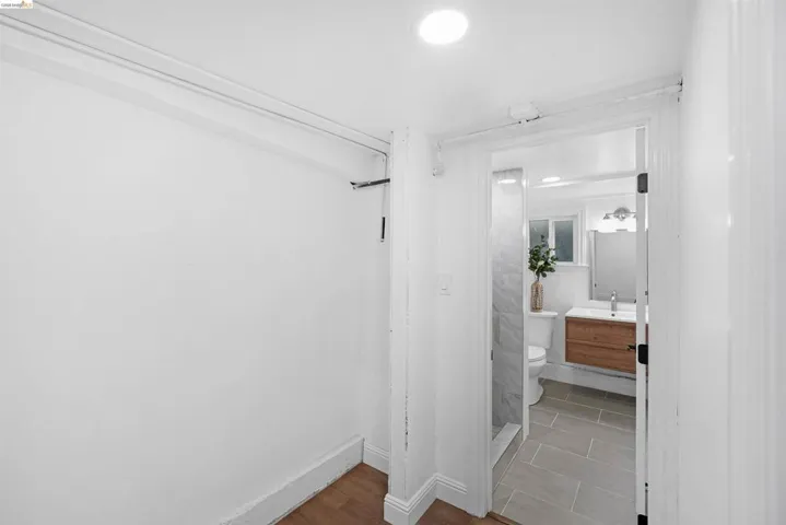 White-painted interior with wood-finish flooring, opening to a bathroom featuring a wood vanity with a white countertop, a shower with stone-look tile, and grey tile flooring