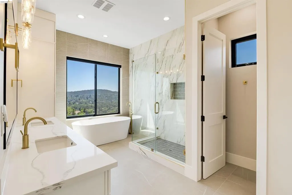 Freestanding soaking tub with a large picture window, dual vanity with undermount sinks and gold-tone fixtures, glass-enclosed shower with tile surround, recessed lighting, and light-toned floor tiling