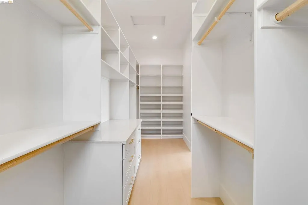 Spacious walk-in closet featuring extensive built-in shelving, hanging rods, central cabinetry with drawers, light wood-finish flooring, and recessed lighting